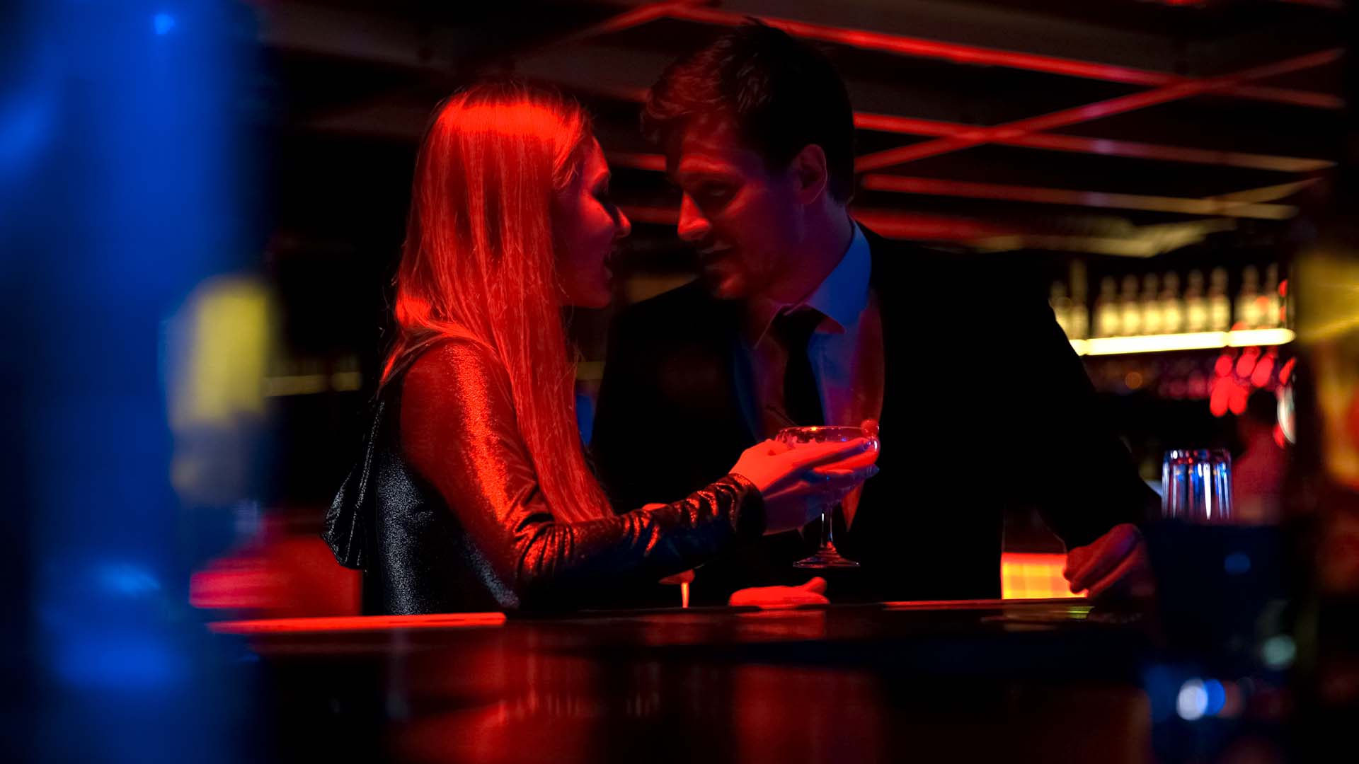 Man in black suit and woman in long-sleeved shirt enjoying drinks while looking at each other. Red colored lights around them.
